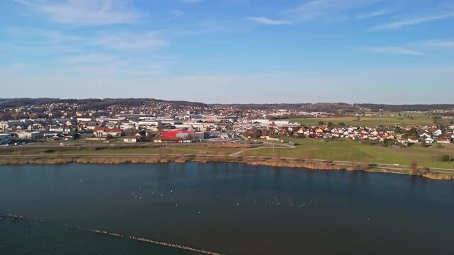 Ptuj, Slovenia and the surrounding Ptujsko Jezero lake are visible from above, showing urban and suburban areas, waterfront, wetlands, farmland, and meadows under a clear sky in an orbital drone view.