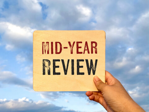 Hand holding a card with mid year review text against sky background, symbolizing business performance evaluation, planning and progress check.