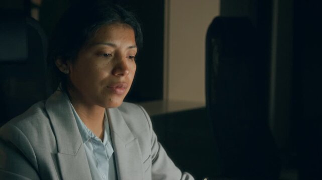 Medium close-up of hands and face of tired Hispanic female accountant working on quarterly report at midnight in darkness in office, staring at screen, rubbing face, suffering from headache