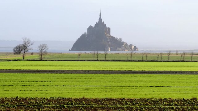 Aerial view of the historic Mont Saint-Michel abbey rising above green agricultural fields under a hazy sky in Mont Saint-Michel, Normandy, France.