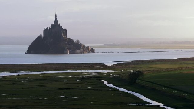 Aerial view of Mont Saint-Michel abbey rising above the tidal bay and green salt marshes under a hazy sky Mont Saint-Michel, Normandy, France.