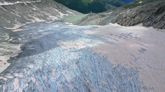 Aerial view of a stunning glacier flowing down the rugged mountain terrain towards a serene lake, creating a landscape of stark beauty, Obergoms, Switzerland.