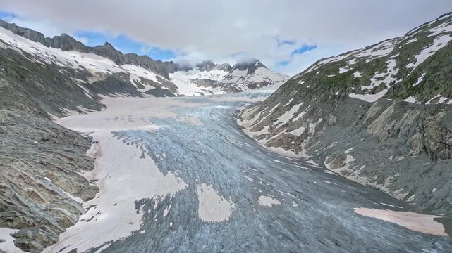 Aerial view of a vast glacier snaking through the rugged terrain, with snow-dusted mountain peaks under a cloudy sky, Obergoms, Valais, Switzerland.