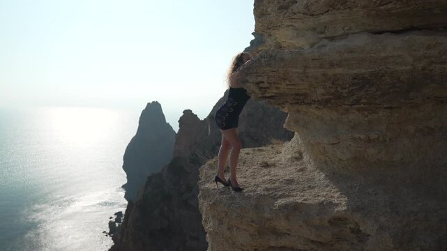 Woman, Cliff, Risk. Woman in a short dress and high heels standing dangerously close to a cliff edge overlooking the sparkling blue sea.