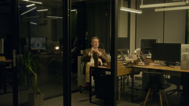 Wide shot of mature Caucasian male corporate manager working on financial report at night in dark office, taking break and napping with feet on desk, computer screen reflected in glass windows