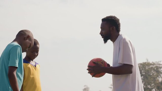 Black coach gives basketball guidance closeup, focused conversation with two black boys on sunlit field, gentle instruction and passing demonstration, encouraging mentorship during outdoor practice