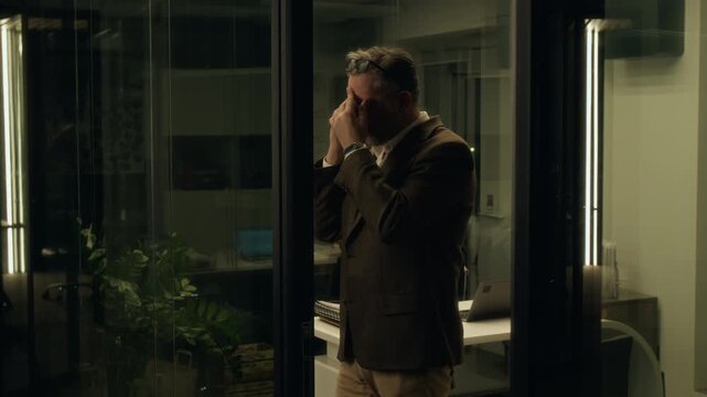 Medium full shot of stressed Caucasian corporate CEO standing in office door, having business conversation on mobile phone, while taking off glasses, fidgeting with jacket, suffering from burnout