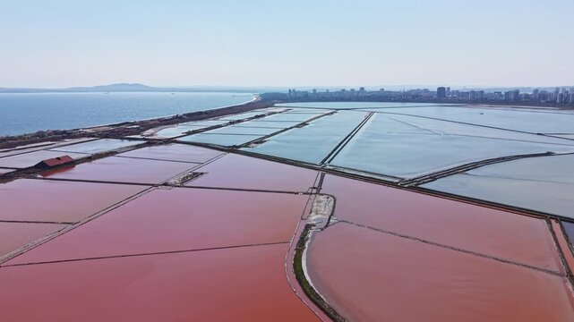 Salt flats stretch out near a coastal city with many rectangular ponds. Different colors appear in the water as sunlight shines on the flats. The distant skyline adds to the view.