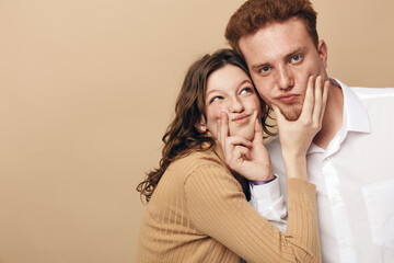 Naklejka premium Young couple with playful expressions posing together on beige background. Woman in brown sweater and man in white shirt show fun and affection. Concept of friendship and joy.
