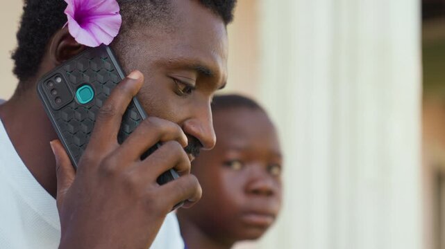Black man on phone with purple flower, young boy watching behind, outdoor veranda portrait, thoughtful expression, warm afternoon light, textured phone case, close profile, community connection,