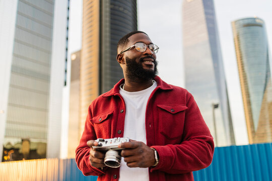 Fashionable man in red jacket holding camera in city environment