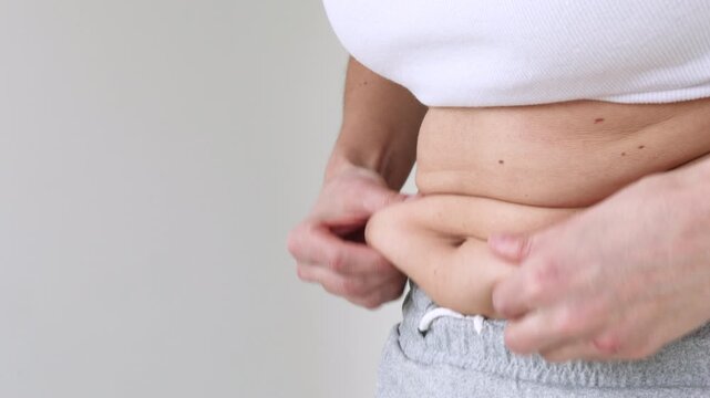 Close-Up of Woman's Hands Pinching Belly Fat Skin Fold at Waist in White Crop Top and Gray skirt &mdash; Female Checking Abdominal Subcutaneous Fat, Body Weight Awareness, Healthy Lifestyle Motivation