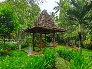 Obraz premium Traditional Javanese Gazebo in Lush Tropical Garden with Stone Pathway, Green Foliage, and Rustic Clay Roof Pavilion in Serene Natural Landscape
