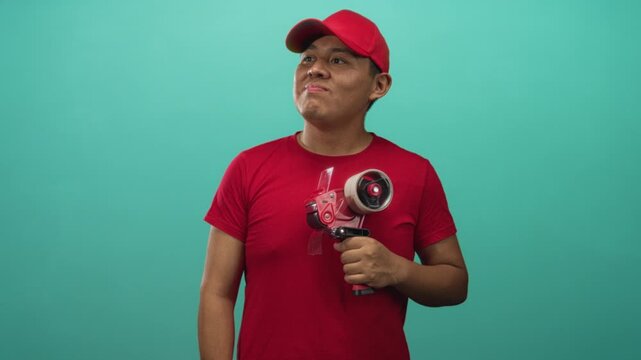 Young hispanic man holding packing tape dispenser in studio with teal backdrop; confident and hardworking.