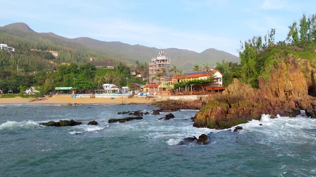 Aerial view of the waves crashing against the rocky shore of Bai Xep Beach, with lush green hills and buildings in the background, Bai Xep, Binh Dinh, Vietnam.