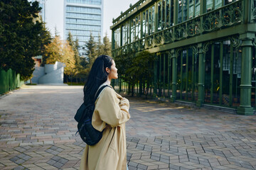 Fototapeta premium woman with backpack wearing a beige coat stands on a tree lined street admiring ornate green architecture in a calm urban setting during autumn, conveying exploration, style, and quiet elegance