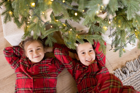 Siblings in red pajamas smiling under Christmas tree at home