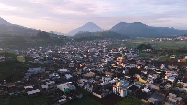 Aerial view of buildings and houses nestled among rolling mountains and volcanoes, showcasing a vibrant cityscape against the natural landscape, Dieng, Central Java, Indonesia.