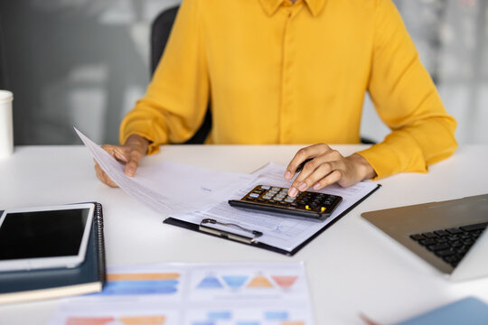 Hands of a person wearing a yellow shirt using a calculator to perform financial calculations while reviewing documents, and preparing taxes on a modern office desk with a laptop and tablet