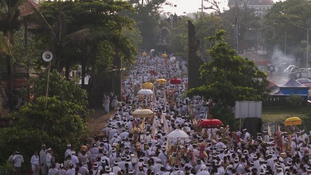Seminyak, Indonesia - 21 March 2026: Aerial view of a large procession of people dressed in white with colorful umbrellas during the day.