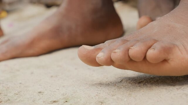 Bare feet on sand, black boy closeup of toes and soles resting on warm textured ground, sandy skin, rural setting, quiet pause before children play, natural light highlighting dust and detail