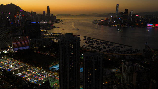 Hong Kong - 14 February 2026: Aerial view of Victoria Harbour at sunset with illuminated skyscrapers, a bustling night market, and boats in the water.