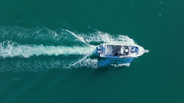 Drone shot of white fishing boat cruising through calm turquoise sea waters leaving foamy wake trail behind. Twin outboard motors power vessel across open ocean on sunny day, top down perspective