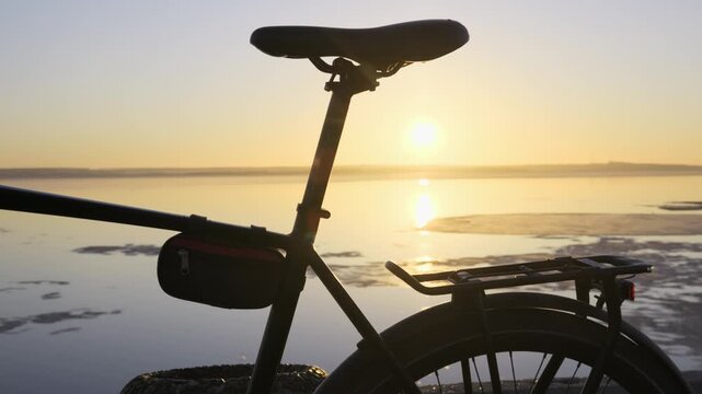 A bicycle silhouette stands parked beside a partially frozen lake during a bright golden sunrise. Concept of peaceful outdoor recreation, winter cycling, and enjoying natural tranquility.