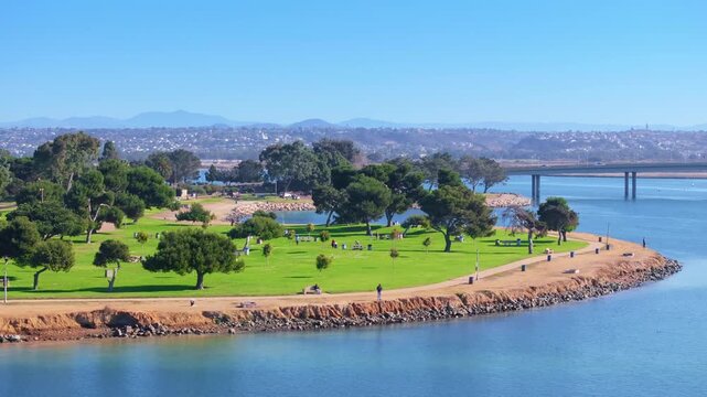 Aerial view of Mission Bay Park island in San Diego featuring lush green grass, scattered trees, walking paths along rocky shoreline, calm blue water, bridge crossing bay