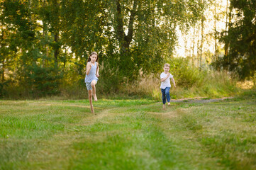 Fototapeta premium Happy Siblings Running Barefoot on Green Grass in Summer Park. Childhood Freedom and Joy Concept. Little Boy and Girl Racing on Meadow at Sunset Golden Hour. Rural Vacation, Active Lifestyle
