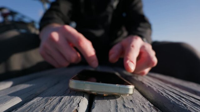 Unrecognizable male hands tap rapidly on a smartphone resting on an outdoor wooden bench. Concept of modern mobile gaming, constant digital communication, and internet addiction.