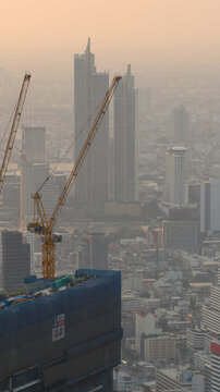 Bangkok, Thailand - 05 February 2026: Aerial view of construction cranes atop a skyscraper under development with workers visible on the roof against the hazy city skyline and high-rise buildings.