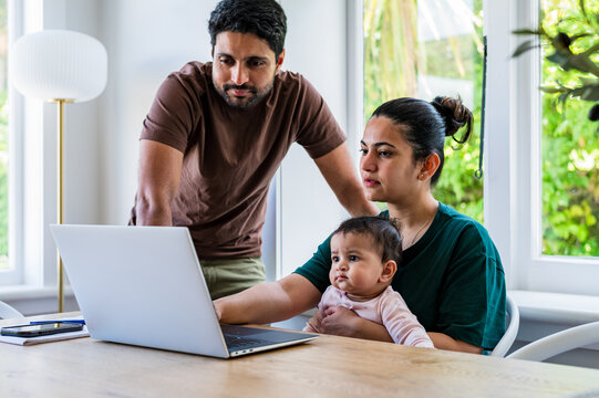 Indian mum working on her laptop with baby girl sits on her lap while husband hovers behind