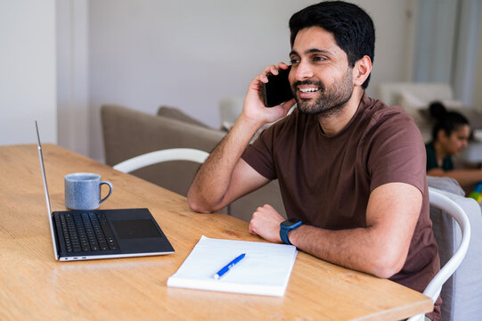 Indian man on a phone call beside a laptop and notebook in a home office space
