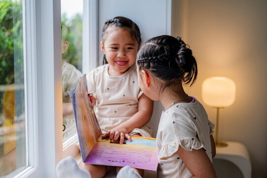 Young girl sitting on the windowsill sharing a storybook with older sister