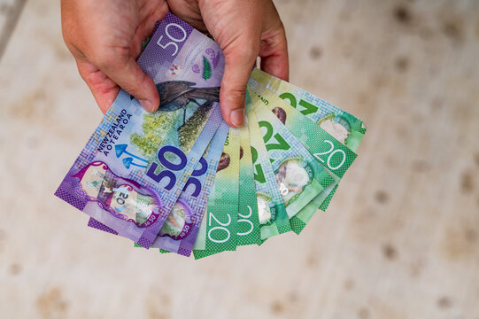 Hands display a mix of New Zealand banknotes including twenty and fifty dollar notes