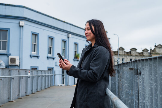 Woman standing on pedestrian bridge while checking smartphone near blue building in New Zealand