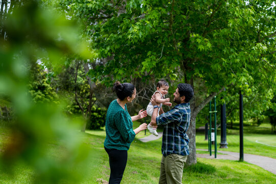 Man lifting a smiling baby as a woman standing close while enjoying a sunny day in a park