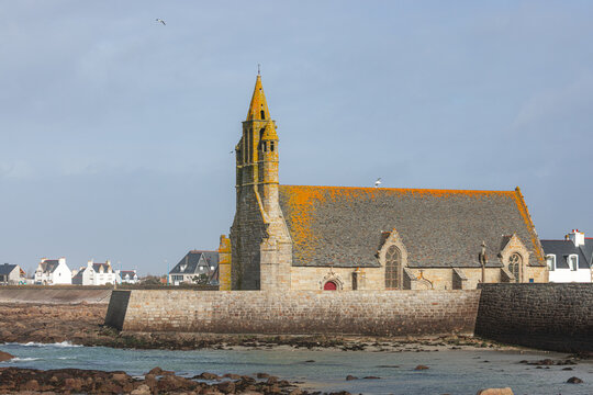 View of the historic stone Chapelle Saint-Pierre with its pointed spire and orange lichen-covered roof standing behind a sea wall on the rocky coast in Penmarch, Bretagne, France.