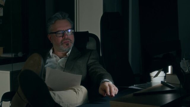 Medium shot of overworked mature Caucasian male corporate lawyer sitting in dark office with feet up on desk in middle of night, studying document draft then typing on computer to make corrections