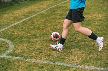 Obraz premium Young athlete kicks soccer ball on grass field. Grass is green with faded white lines. Player wears blue shirt and black shorts. Shoes are white with colorful accents. Game appears to be in progress