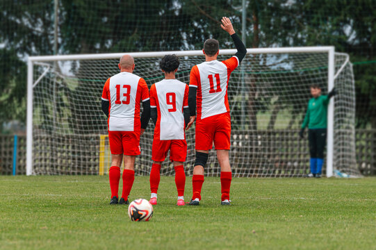 Three soccer players in red and white kits standing on a grass field, depicting a local village football match