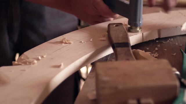 Woodworking workshop scene with a metal clamp holding curved timber on a messy workbench. Shavings and a mallet in the background. Slow motion sequence.