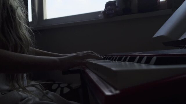 A moody, static side-angle shot capturing a young girl's hands as she plays a red digital piano. The footage focuses on the coordination between both hands on the keys, set in a quiet, indoor environm