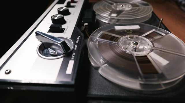 Vintage reel tape machine plays magnetic audio tape. Clear plastic spools spin while reproducing authentic analog sound. The silver control panel highlights classic retro technology.