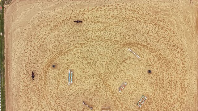 Horses walk and trot in a circular pattern around jumps in a sandy arena during a clear day. The scene shows action and movement in a training setting.