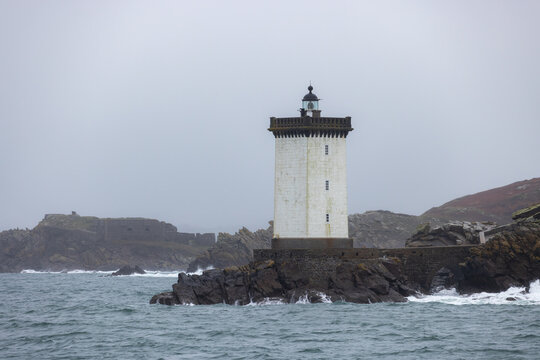 View of Kermorvan Lighthouse standing on a rocky cliff with waves crashing against the shore under a grey overcast sky Le Conquet, Bretagne, France.