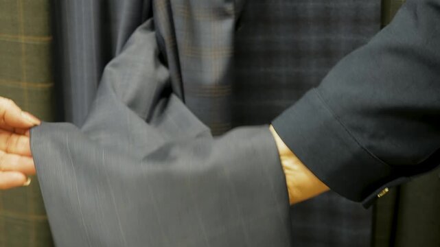 Professional tailor in black uniform displays various high quality checkered wool fabrics including grey and brown patterns for bespoke suit manufacturing in atelier workshop