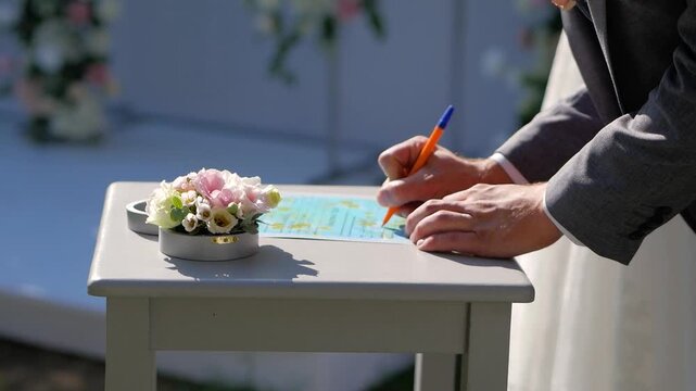 groom hands signing official marriage certificate document followed by bride in white dress picking up pen to sign during outdoor wedding ceremony next to floral arrangement