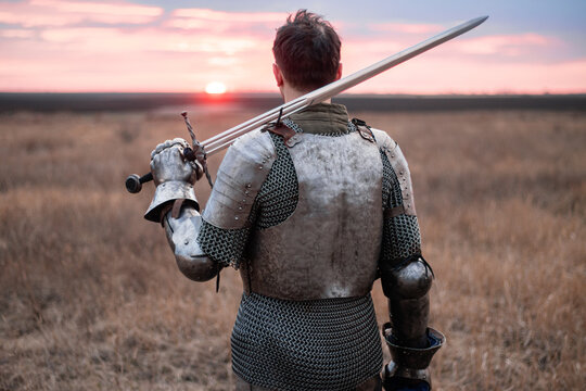 A knight in cuirass and chainmail, holding a sword and a bloodied face, stands with his back to the sunset in a field. A medieval warrior after a battle.